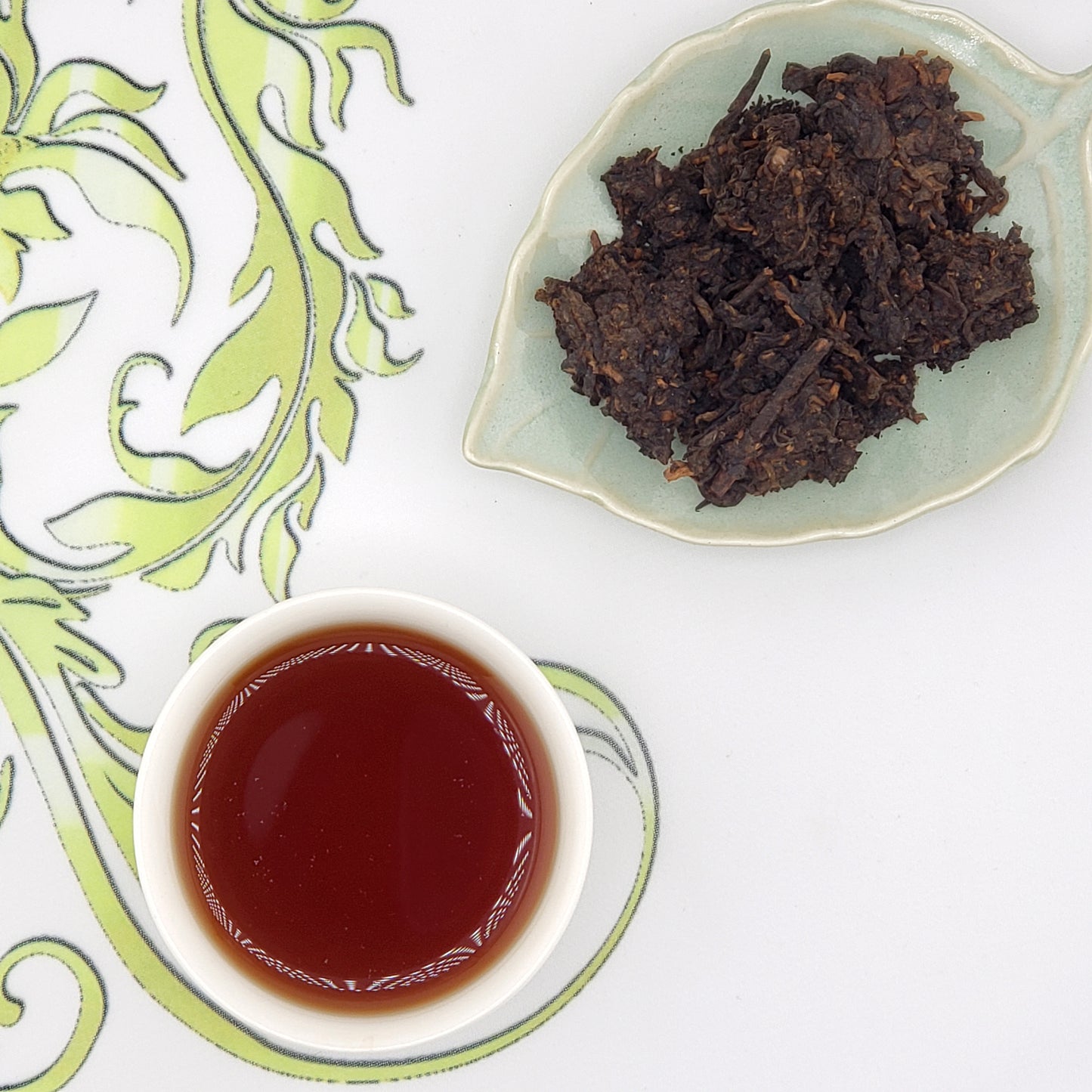 Dried tea leaves on a small plate with a cup of brewed tea on a decorative white and green surface.