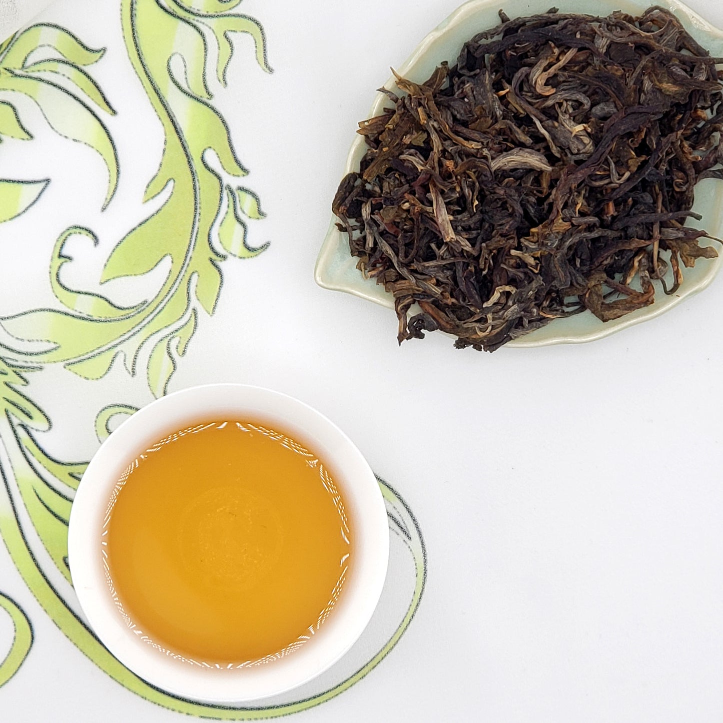 Dried tea leaves on a white plate with a cup of brewed tea on a decorative green and white background.