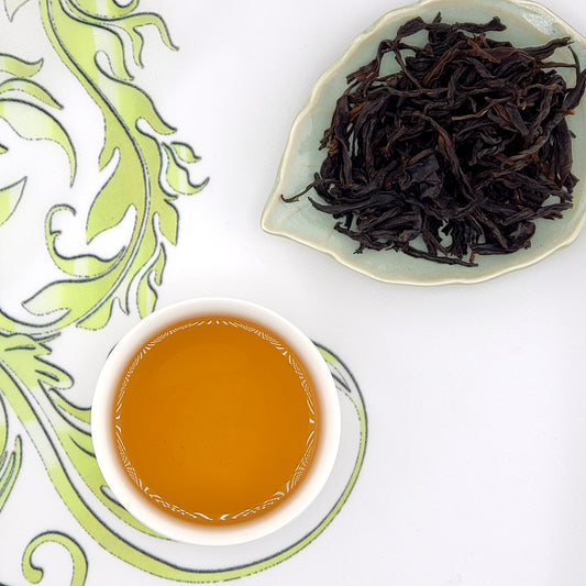 Dried black tea leaves on a leaf-shaped dish with a cup of brewed tea on a decorative white and green plate.