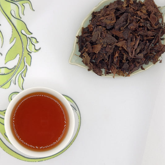 Dried tea leaves on a plate with a cup of brewed tea on a decorative mat.