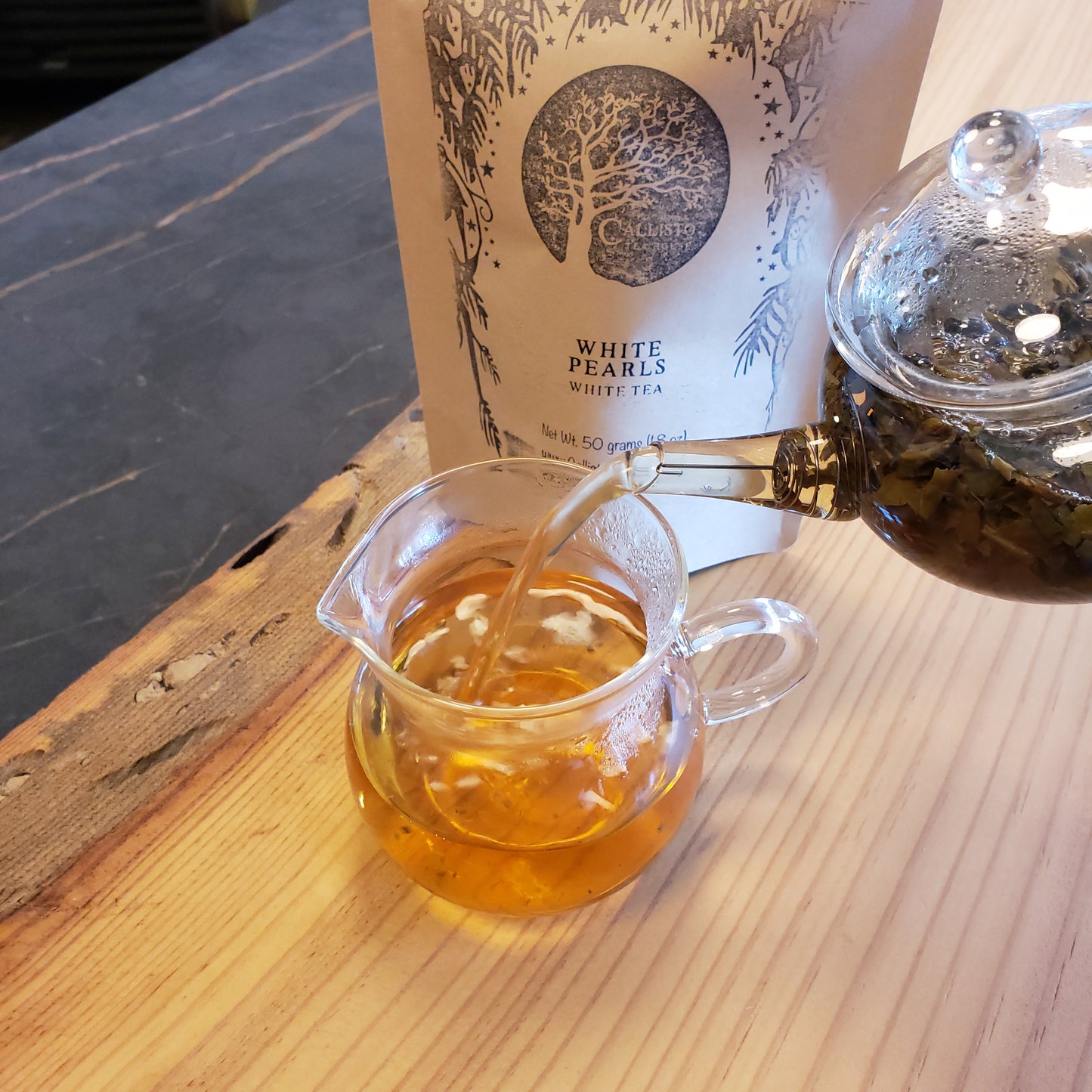 Glass teapot pouring tea into a clear glass mug with a package of White Pearls tea nearby.