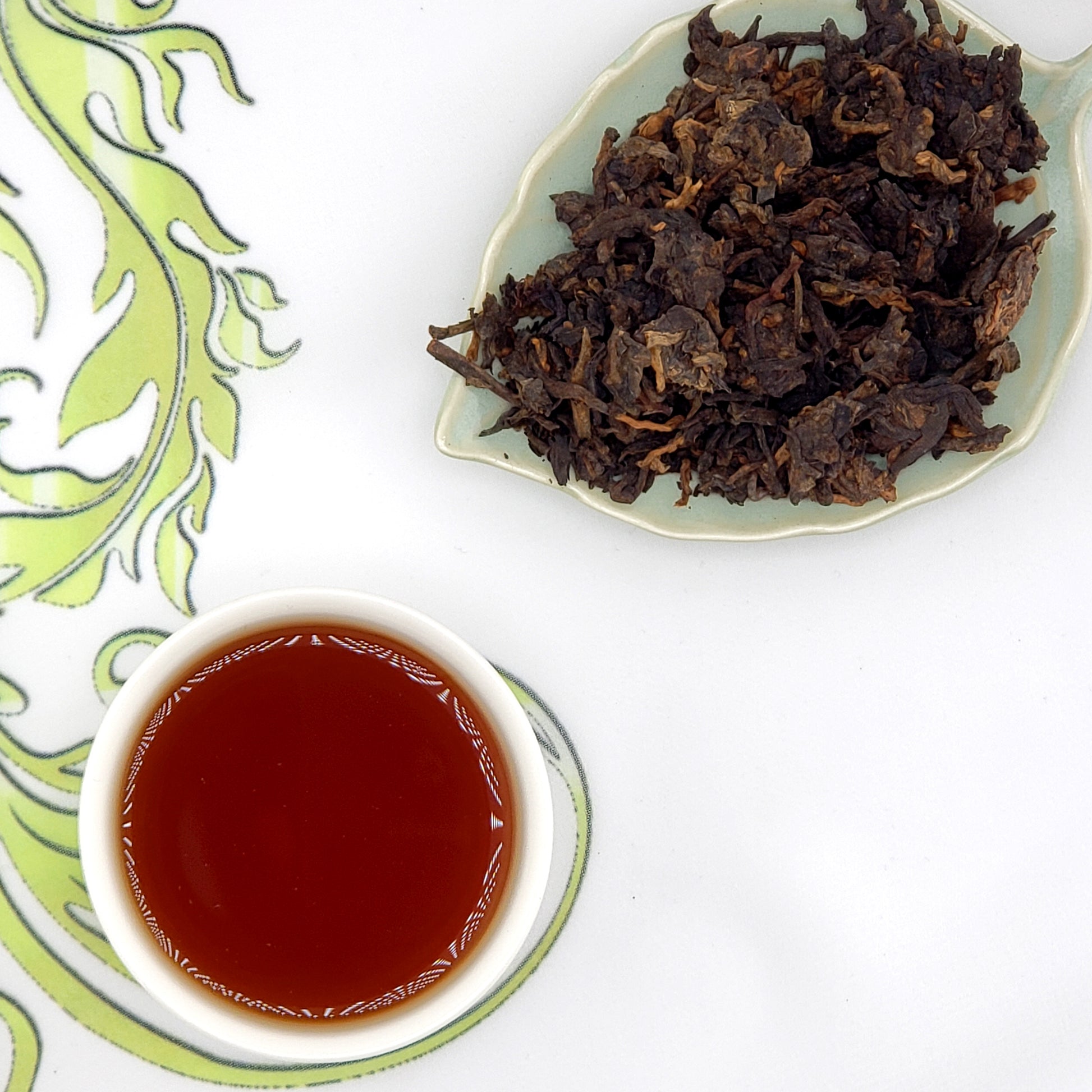 Dried tea leaves on a leaf-shaped plate with a cup of brewed tea on a decorative mat.