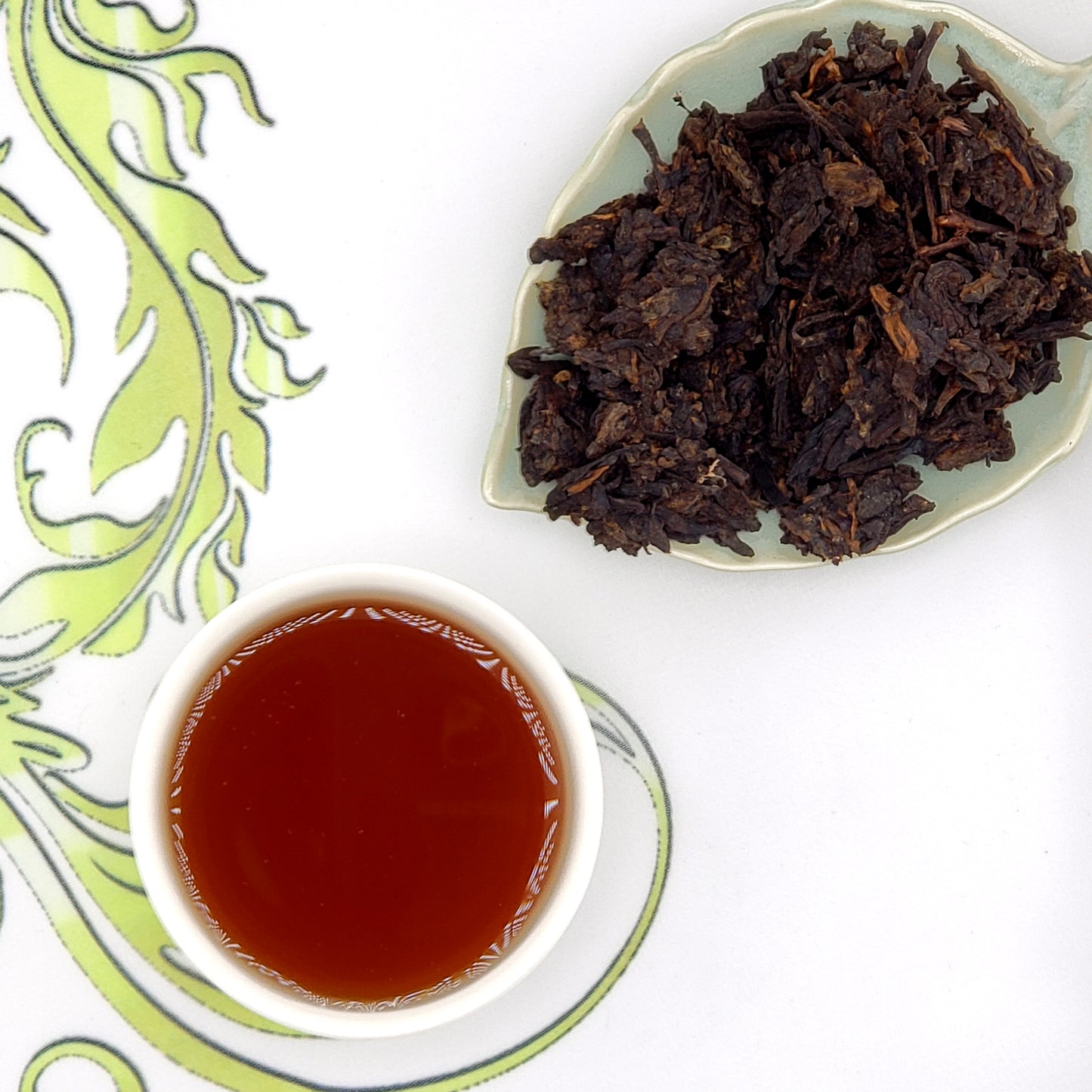 Dried tea leaves on a plate with a cup of brewed tea on a decorative white and green background.