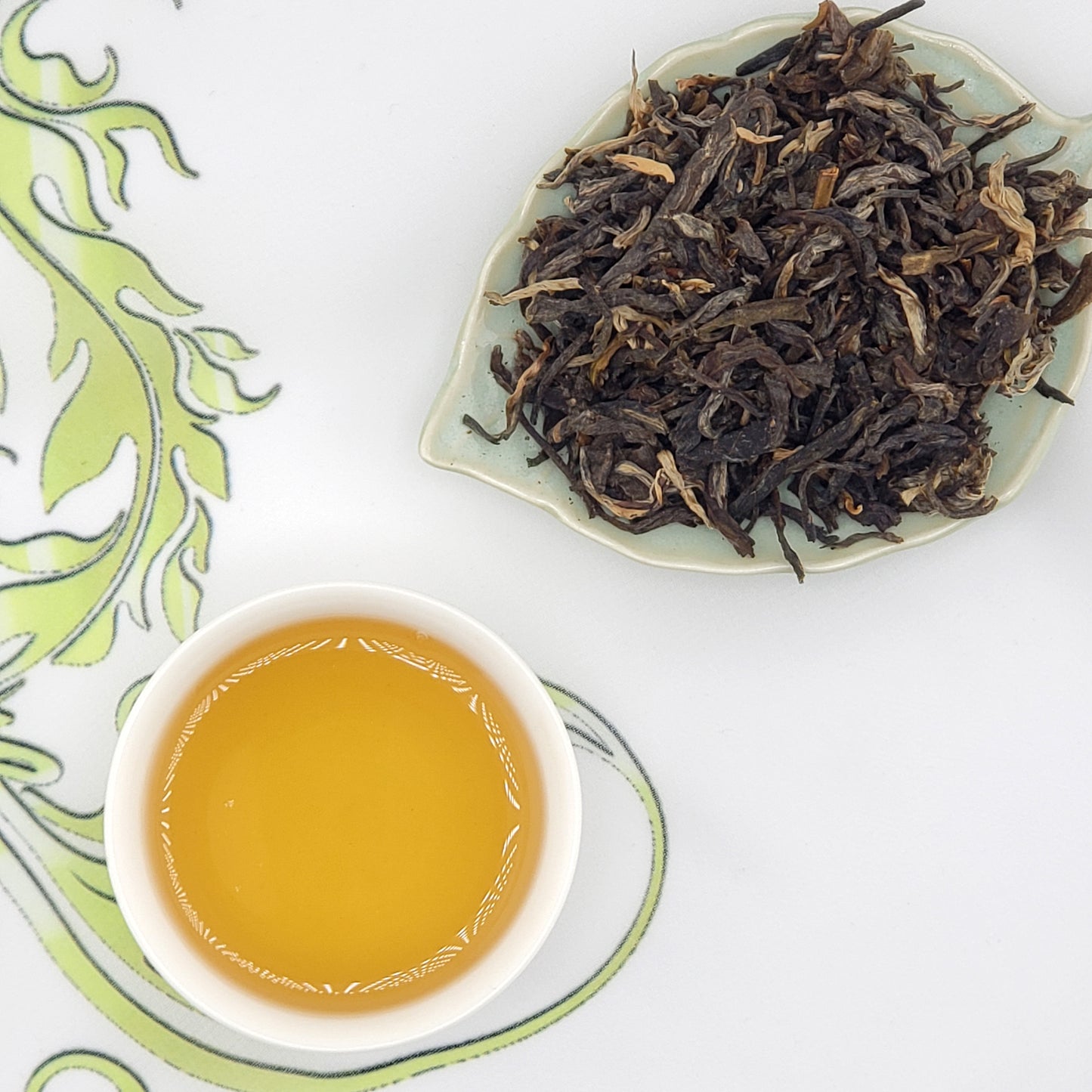 Dry tea leaves on a plate with a cup of brewed tea on a decorative white and green background.