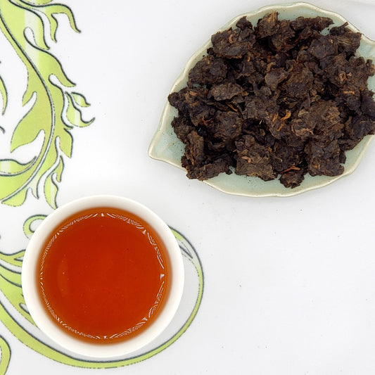 Tea leaves and a cup of light amber colored tea on a decorative tablecloth 