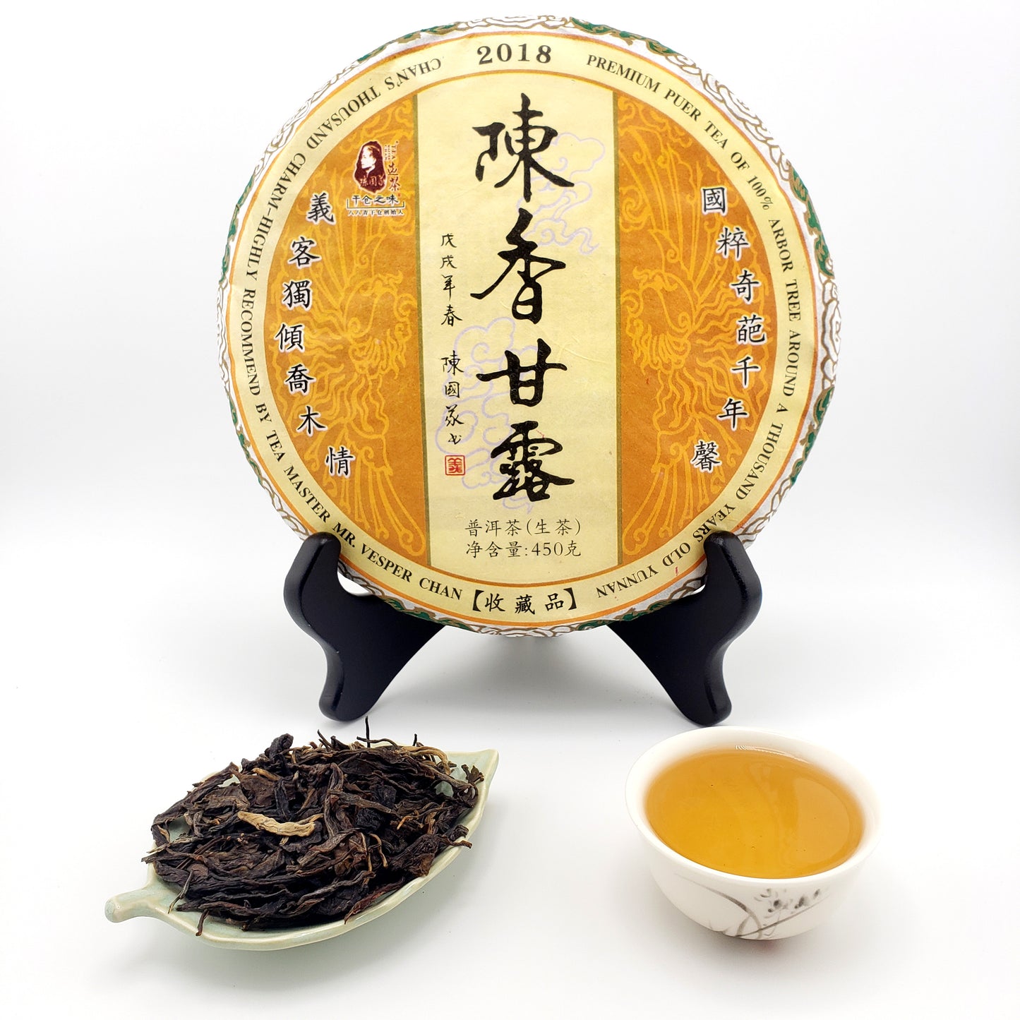 Round tea cake with a yellow label next to a cup of tea and a tea leaf sample on a white background