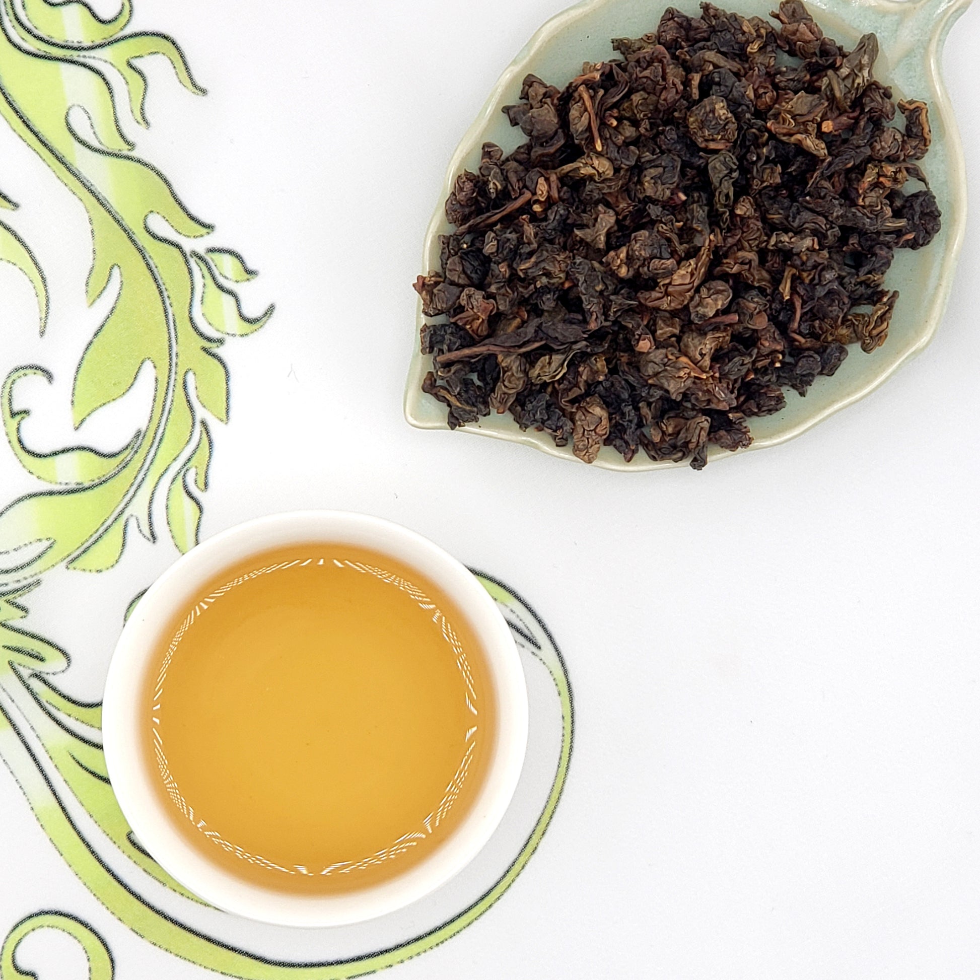 Dry tea leaves on a leaf-shaped plate with a cup of brewed tea on a decorative white background.