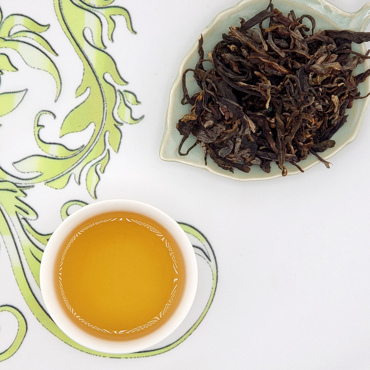 Dried tea leaves on a leaf-shaped dish with a cup of brewed tea on a decorative mat.