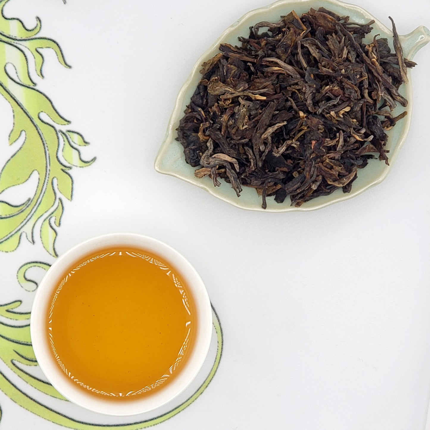 Dried tea leaves on a leaf-shaped plate with a cup of brewed tea on a white background