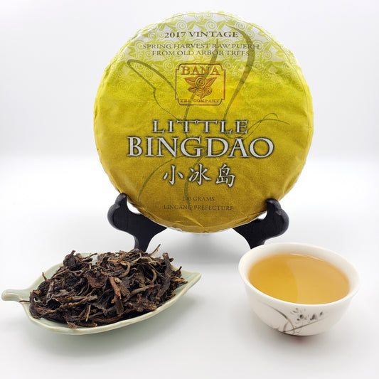 Yellow tea cake labeled 'Little Bingdao' with a teacup of tea and a scoop of tea leaves on a white background.