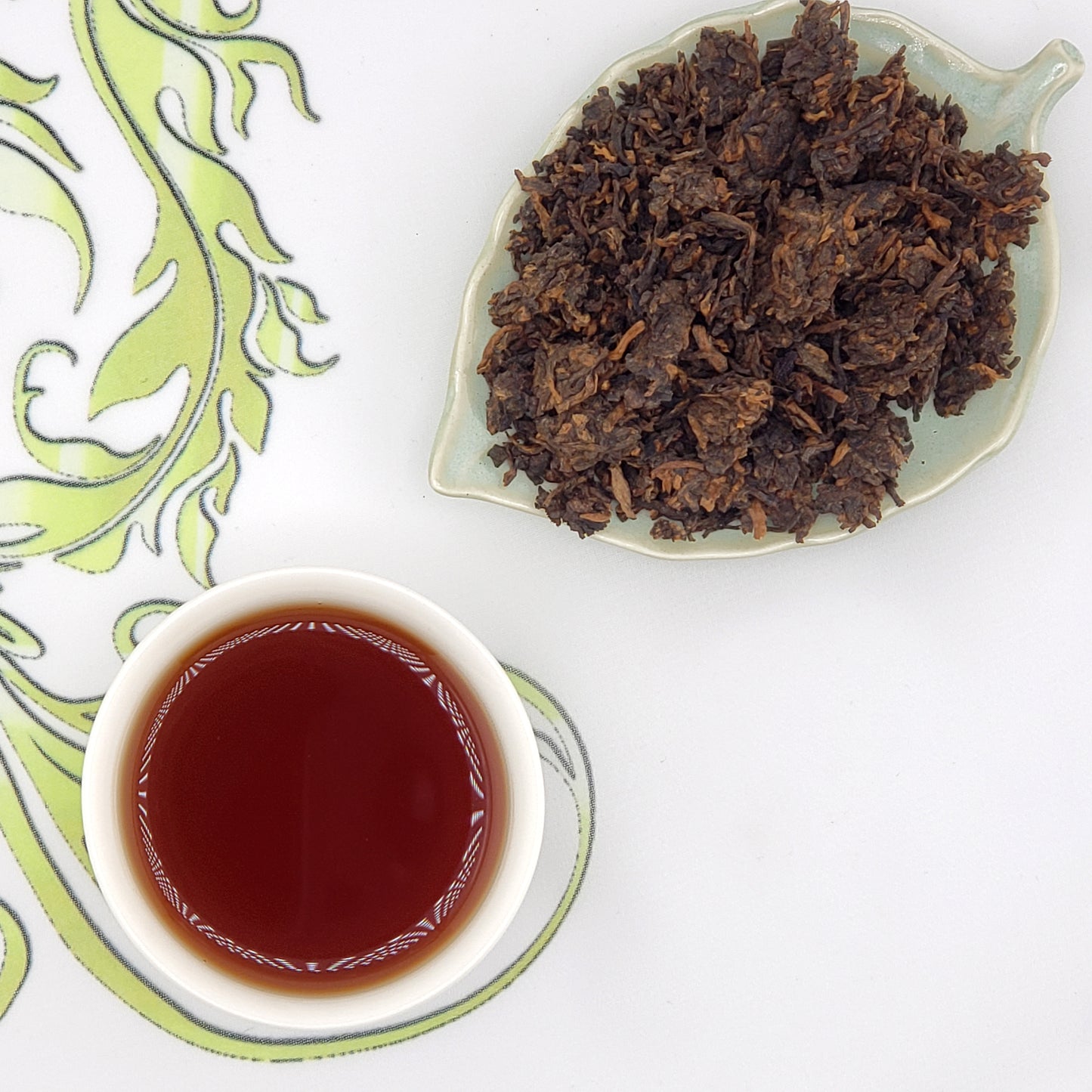 Dried tea leaves on a leaf-shaped plate with a cup of brewed tea on a decorative mat.