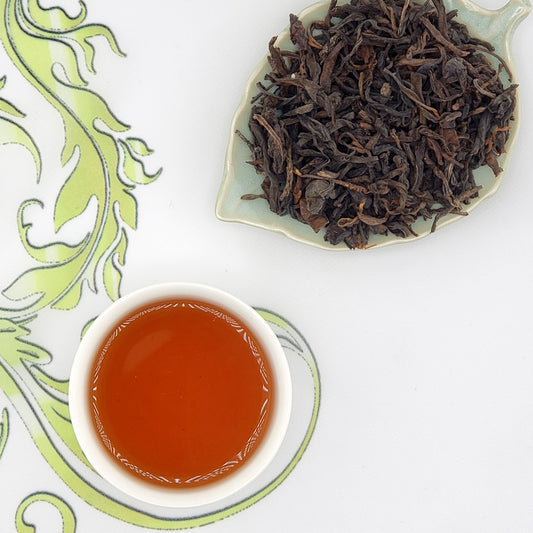 Dried tea leaves on a leaf-shaped plate with a cup of brewed tea on a decorative white and green background.